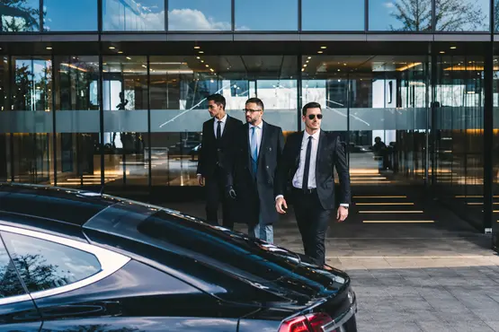 Trois hommes en costume près d’une berline devant un immeuble de bureaux.